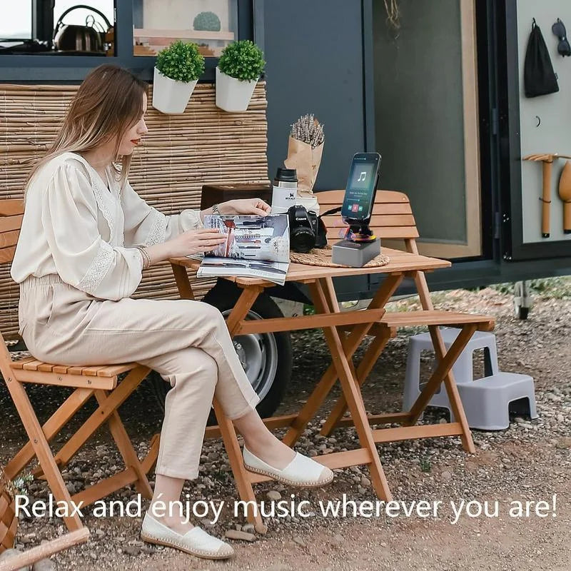 Woman sitting at a wooden table outdoors with a music player and smartphone, surrounded by plants and outdoor furniture.