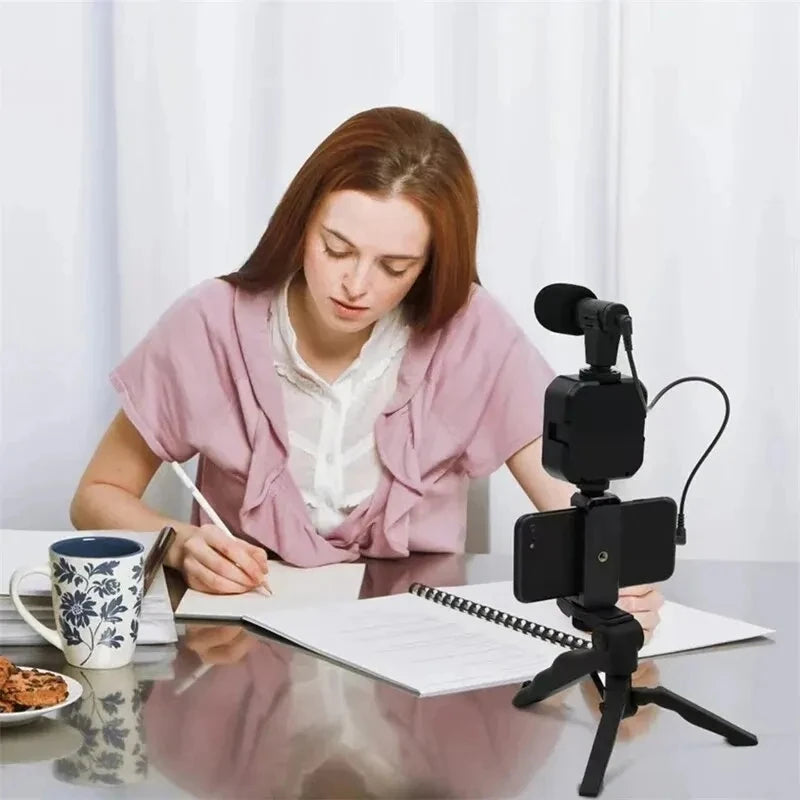 Woman sitting at a table with a smartphone on a tripod, writing in a notebook.